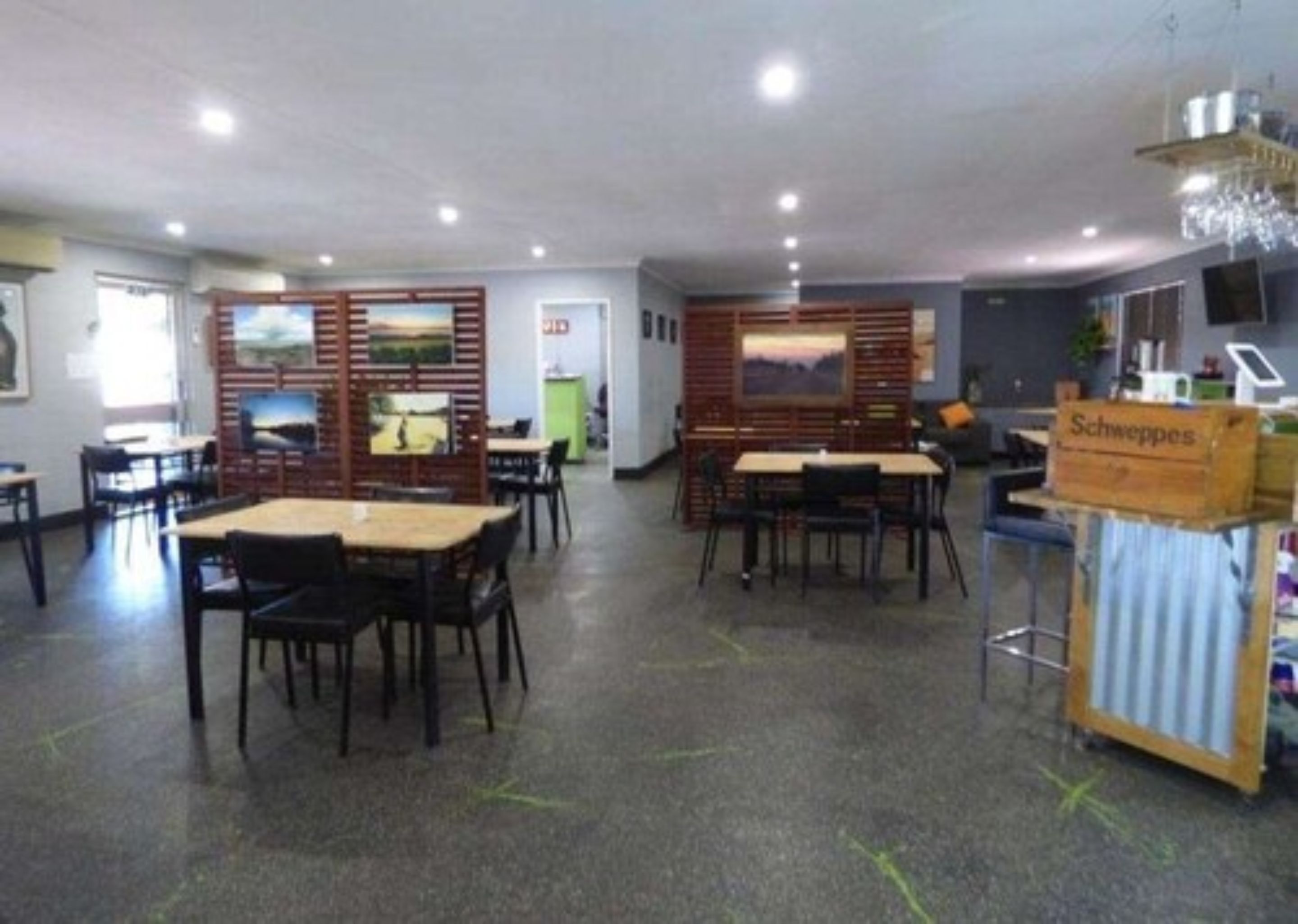Un grand espace dédié à la salle à manger, avec plusieurs tables et chaises, une cloison en bois ornée d’œuvres d’art et un panneau indiquant « Schweppes ».