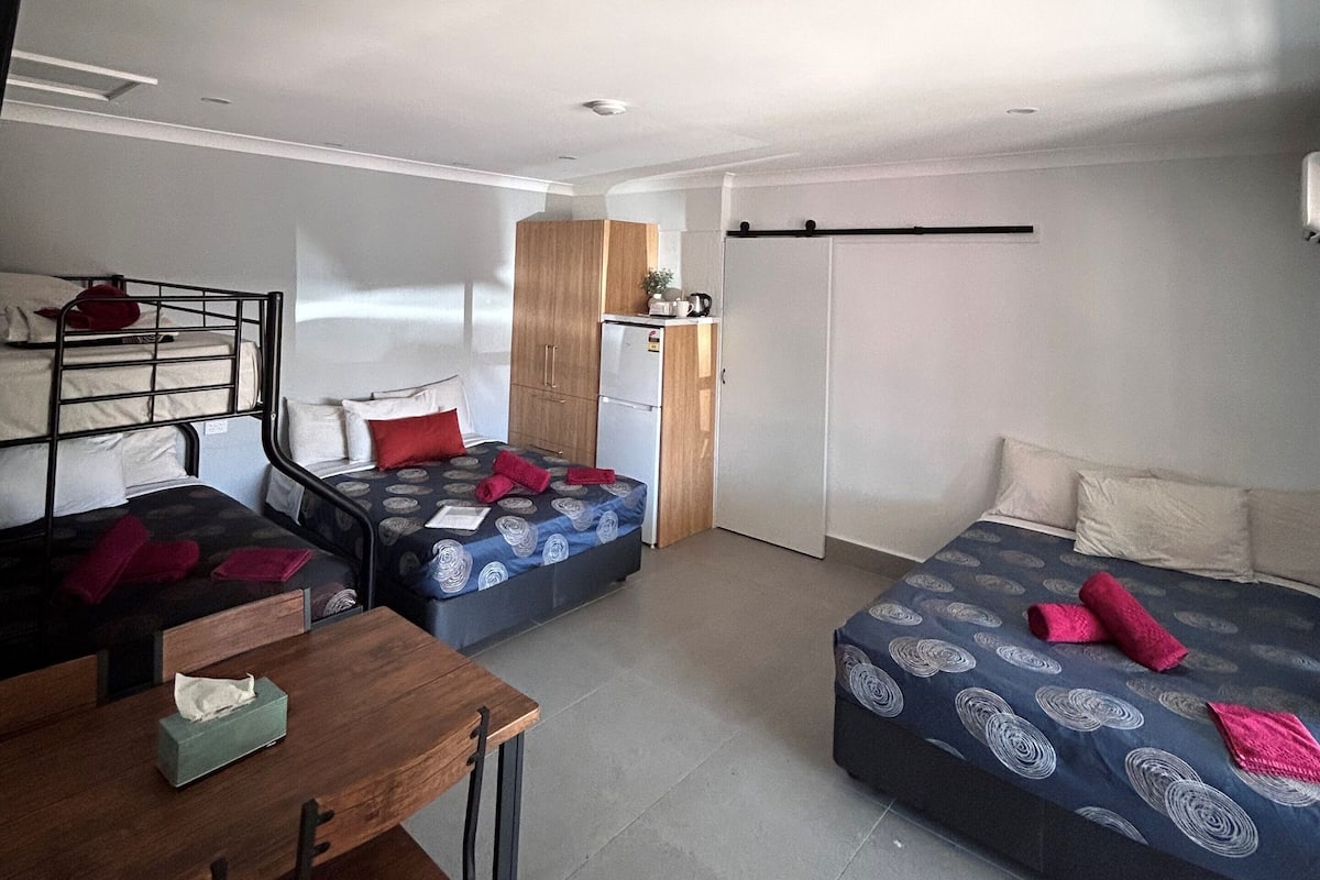 A compact room with two beds, a small kitchenette, a wooden table with chairs, and a ceiling light.