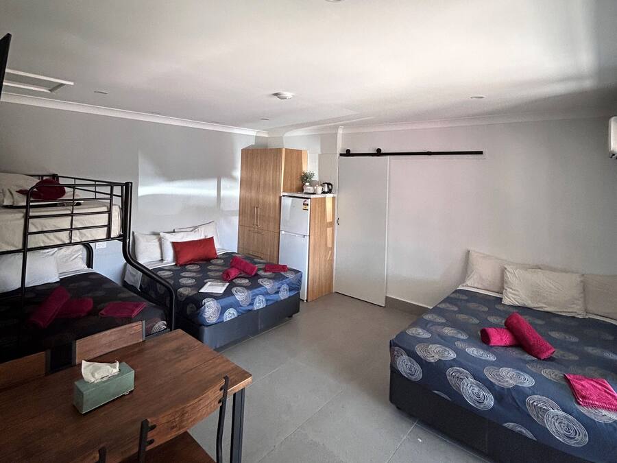A compact room with two beds, a small kitchenette, a wooden table with chairs, and a ceiling light.