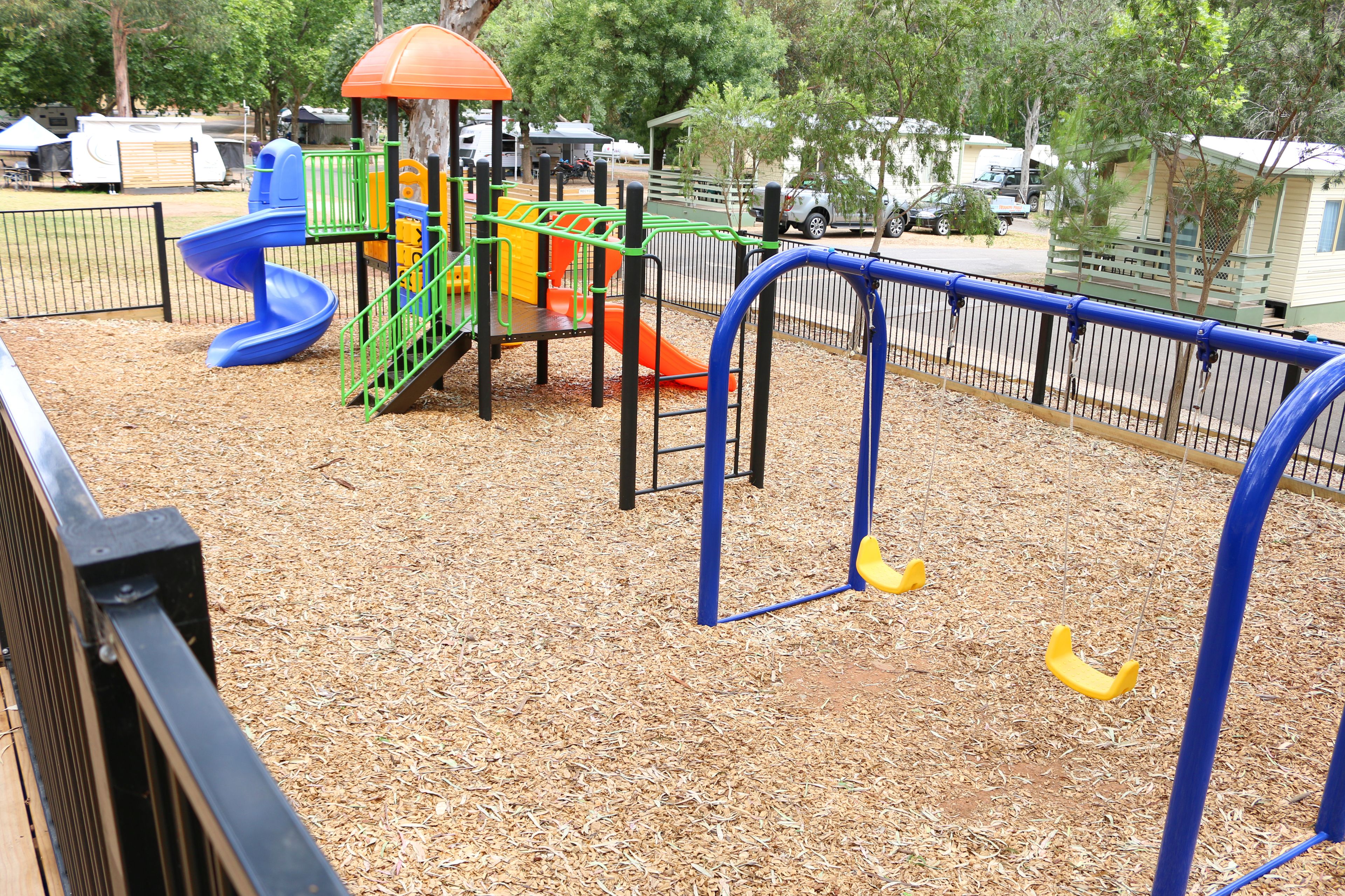 Un parque infantil con un tobogán azul, estructuras de juego naranja y amarillas, y una zona vallada.