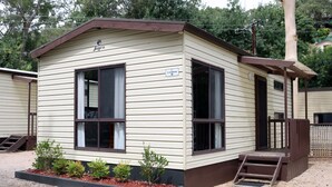 A small, single-story mobile home with a brown roof and beige siding, labeled "Cabin 8." It features two large windows, a small porch with a wooden railing, and a set of steps leading to the entrance.
