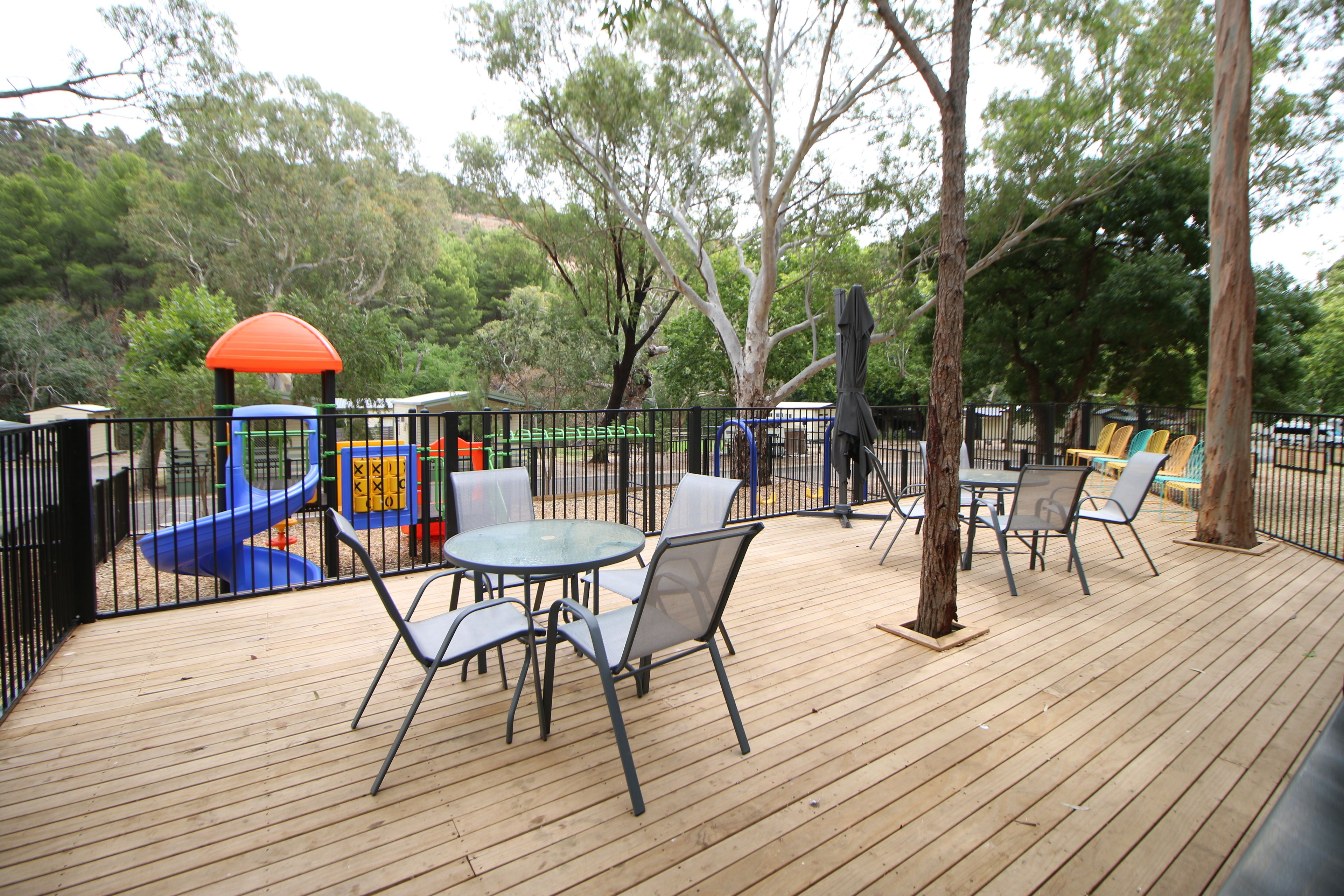 Una terraza de madera con mobiliario exterior y un parque infantil al fondo.