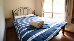 A neatly made bed with striped bedding and a rolled towel, a wooden headboard, and a window with curtains.