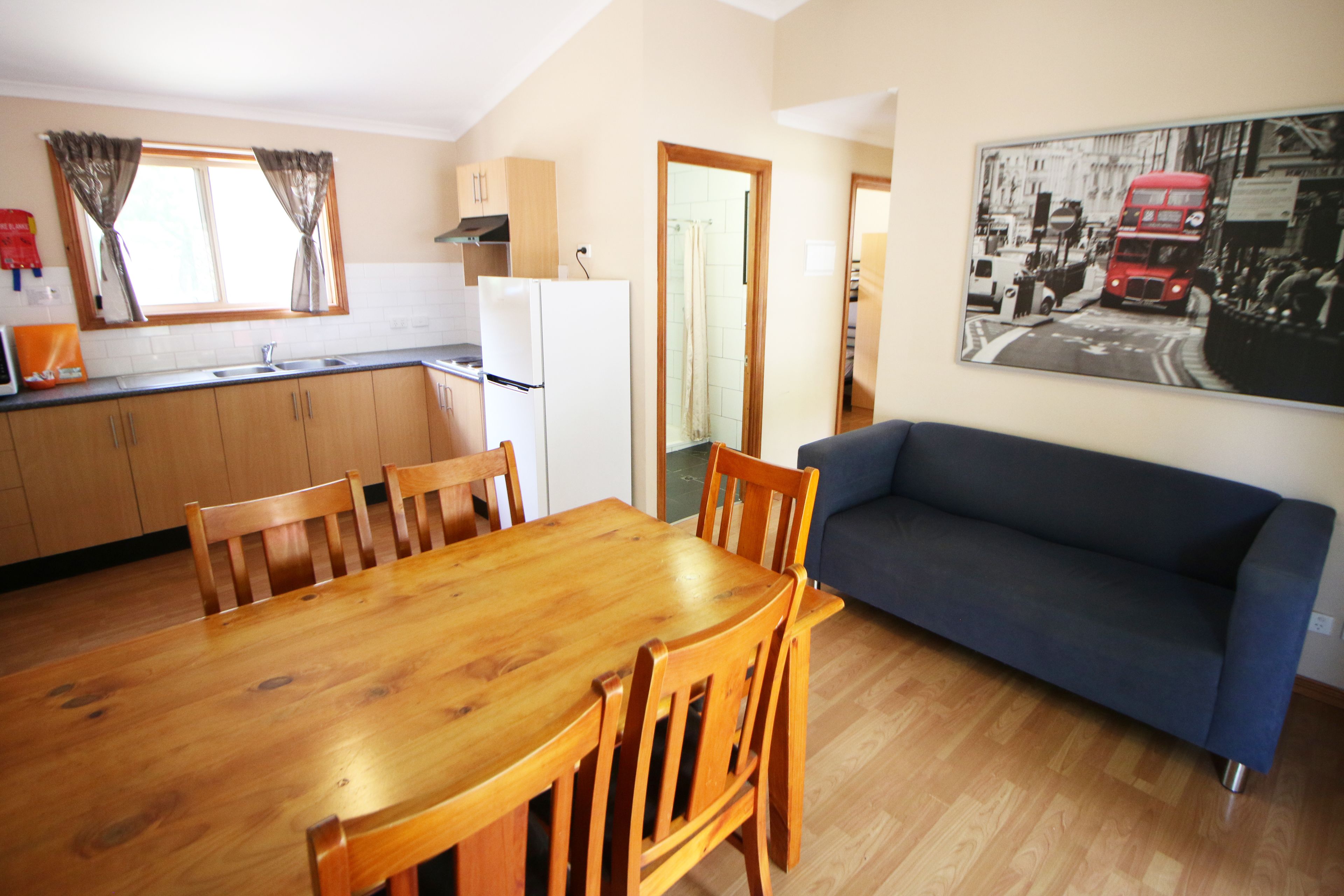 Une cuisine de taille modeste, avec des meubles en bois, un réfrigérateur blanc et une table à manger avec des chaises. Un canapé bleu est placé contre le mur, et un tableau représentant une scène de rue en noir et blanc est visible.