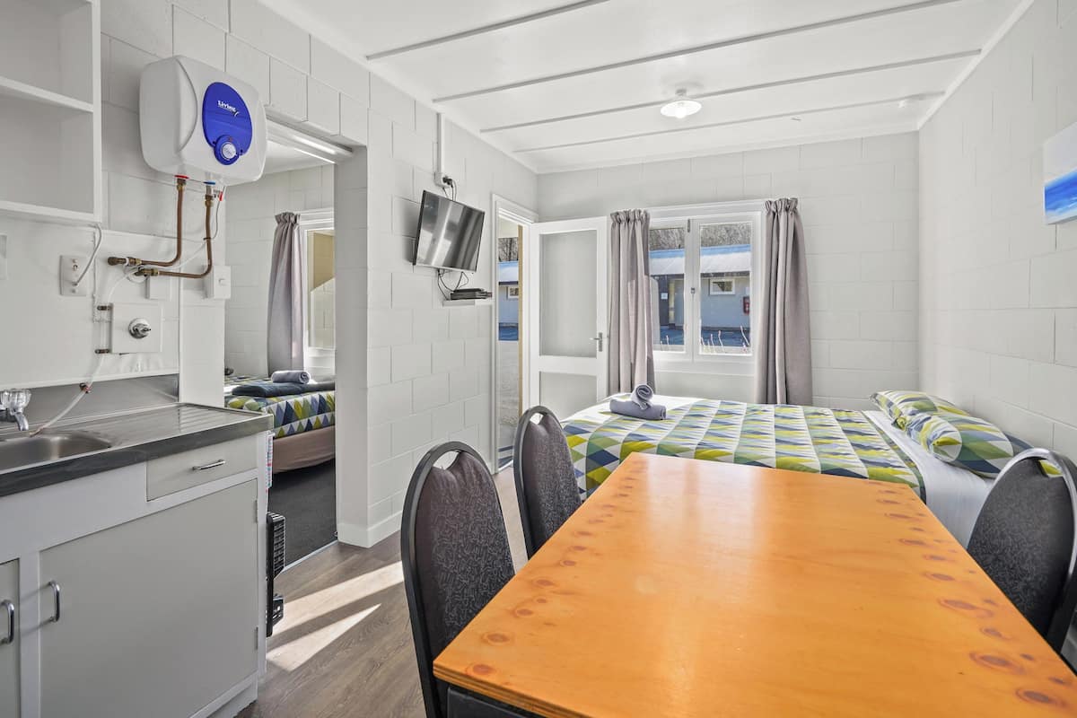 A room with a wooden table, two chairs, a bed with a colorful quilt, a TV mounted on the wall, and a window with curtains.