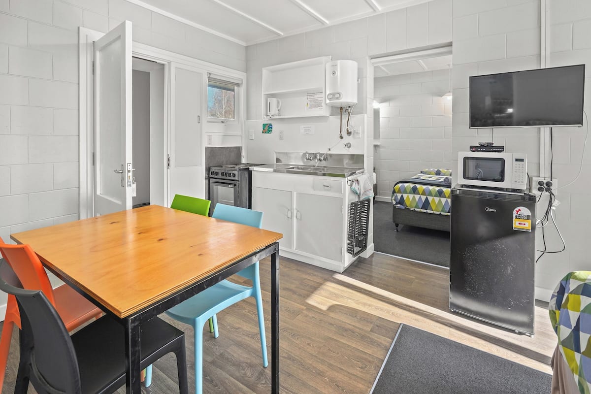 A compact kitchen with a microwave, stove, and sink. There is a dining table with chairs and a TV mounted on the wall.