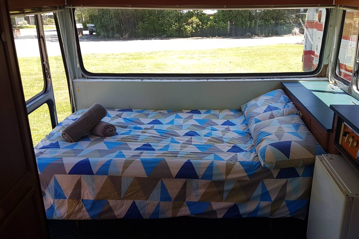 A bed with a geometric patterned bedspread and a rolled towel, a small table with a picture frame, and a window with a view of a grassy area and trees.