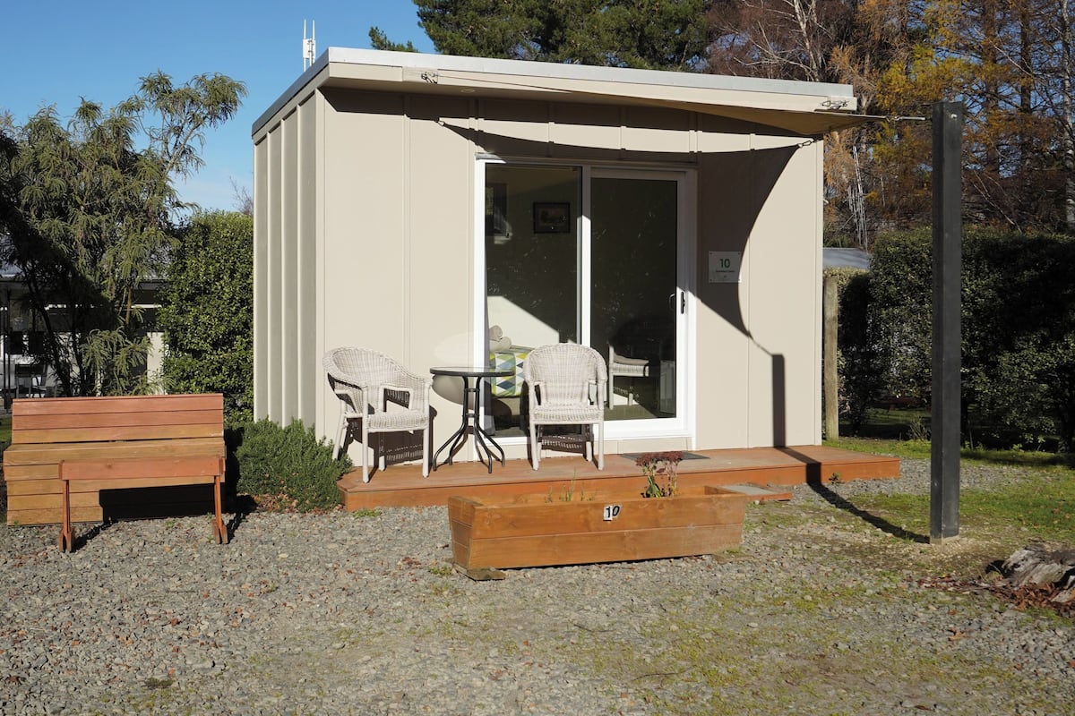 A small, single-story building with a sliding glass door entrance, a wooden bench, two wicker chairs, and a small table.