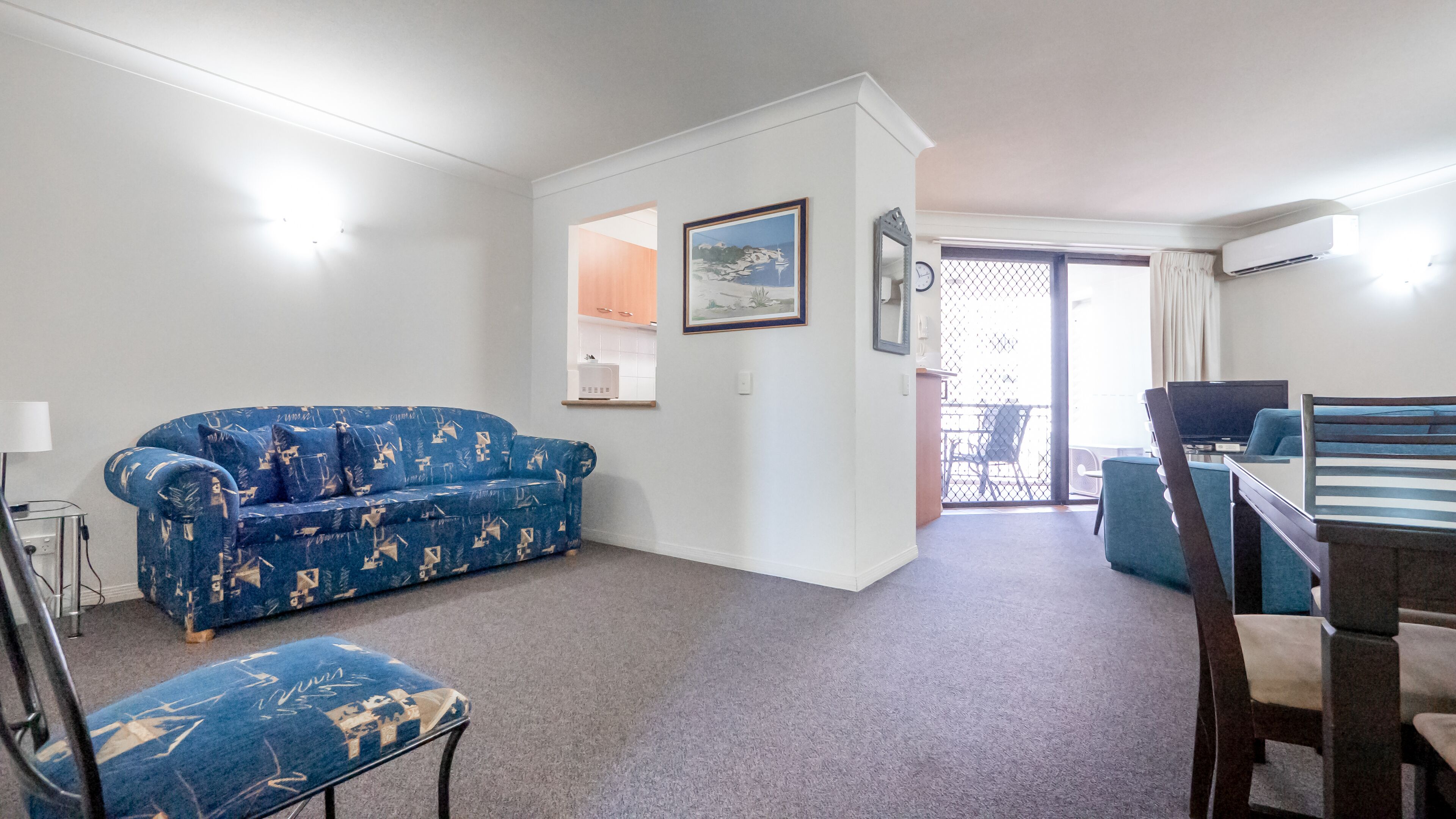 A living room with a blue patterned sofa, a small table, and a chair. There is a door leading to a balcony with a view.
