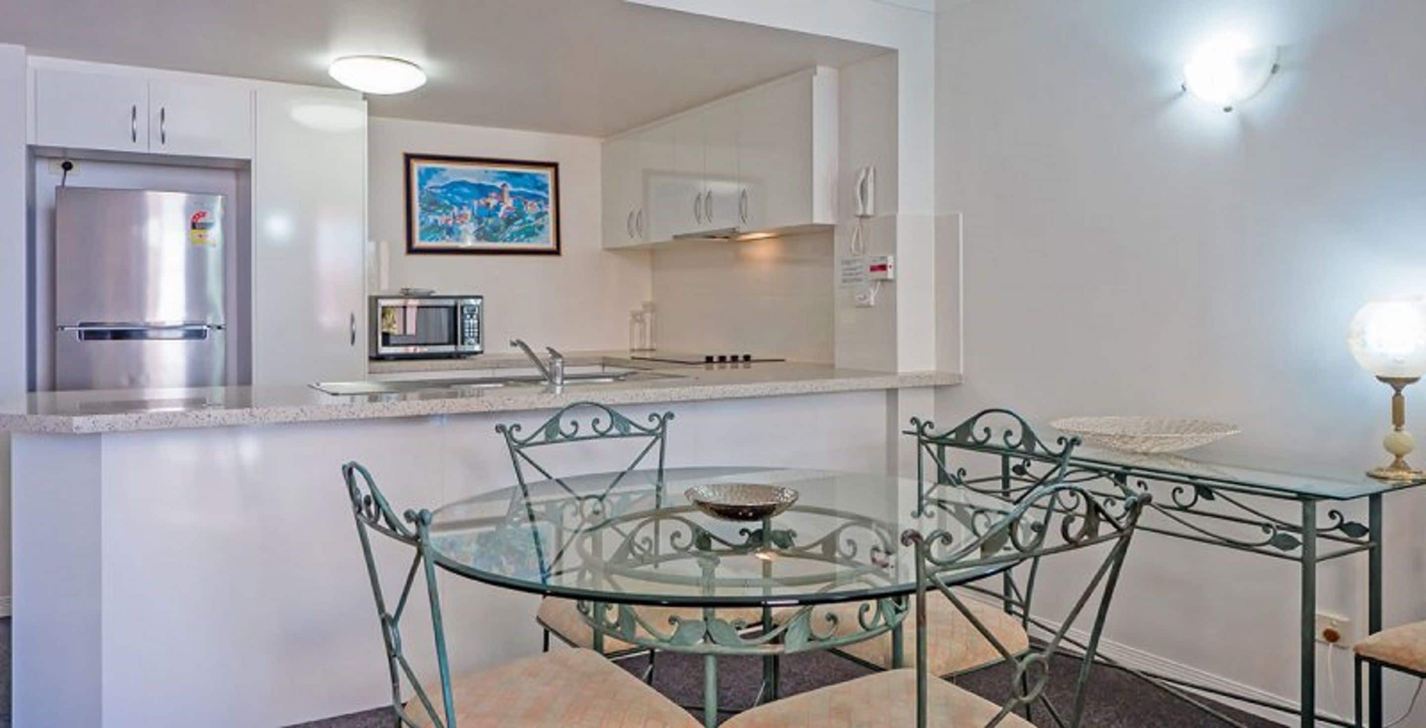 A modern kitchen with white cabinets, a stainless steel refrigerator, a microwave, and a glass dining table with metal chairs.