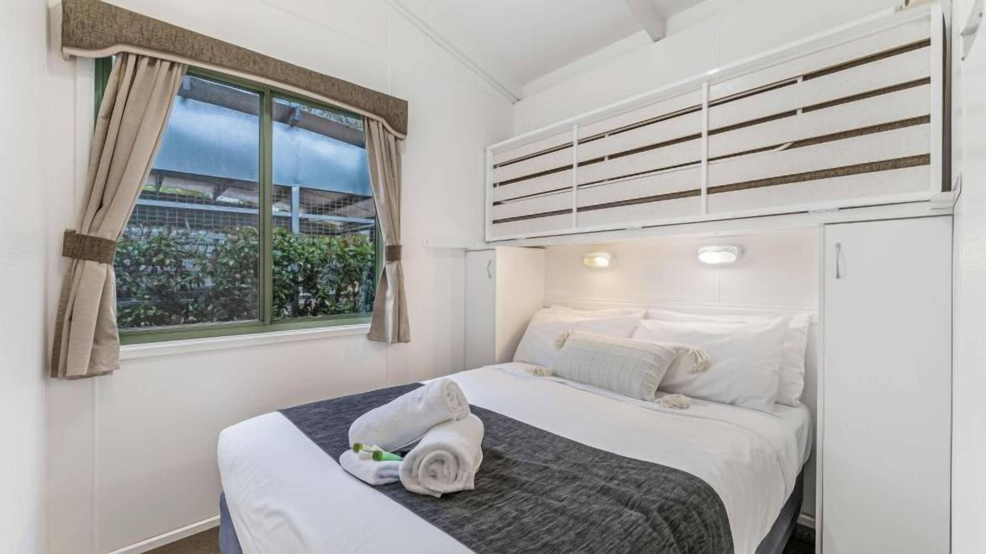 Una cama individual con cabecera gris, ropa de cama blanca y una toalla sobre la cama. Se observa una ventana con cortinas y un equipo de aire acondicionado encima de la cama.