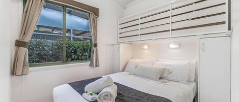 Una cama individual con cabecera gris, ropa de cama blanca y una toalla sobre la cama. Se observa una ventana con cortinas y un equipo de aire acondicionado encima de la cama.