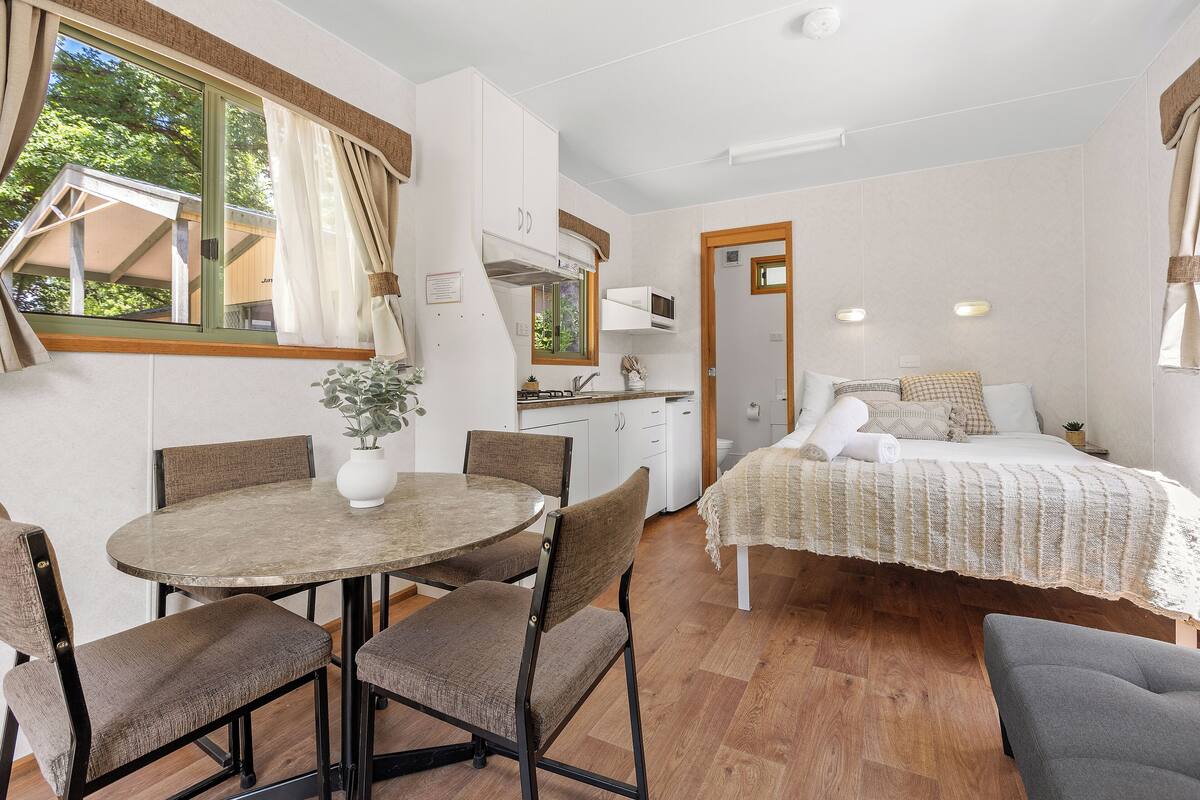 Quarto compacto com mesa de jantar, cadeiras, cama, cozinha compacta e janela com cortinas.
