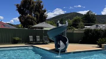 A swimming pool with a water feature, surrounded by a fenced yard with chairs and a tree.