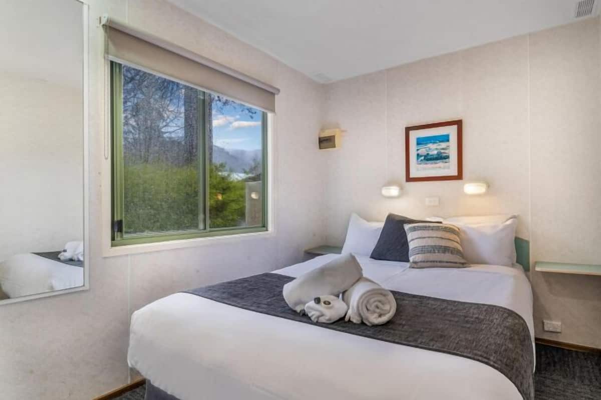 Quarto de hotel com uma cama grande, uma mesinha de cabeceira, um quadro na parede e uma janela com vista para árvores e montanhas.
