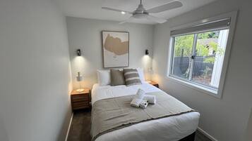 Un dormitorio con cama, ventilador de techo, lámparas de noche, ventana y una obra de arte colgada en la pared.