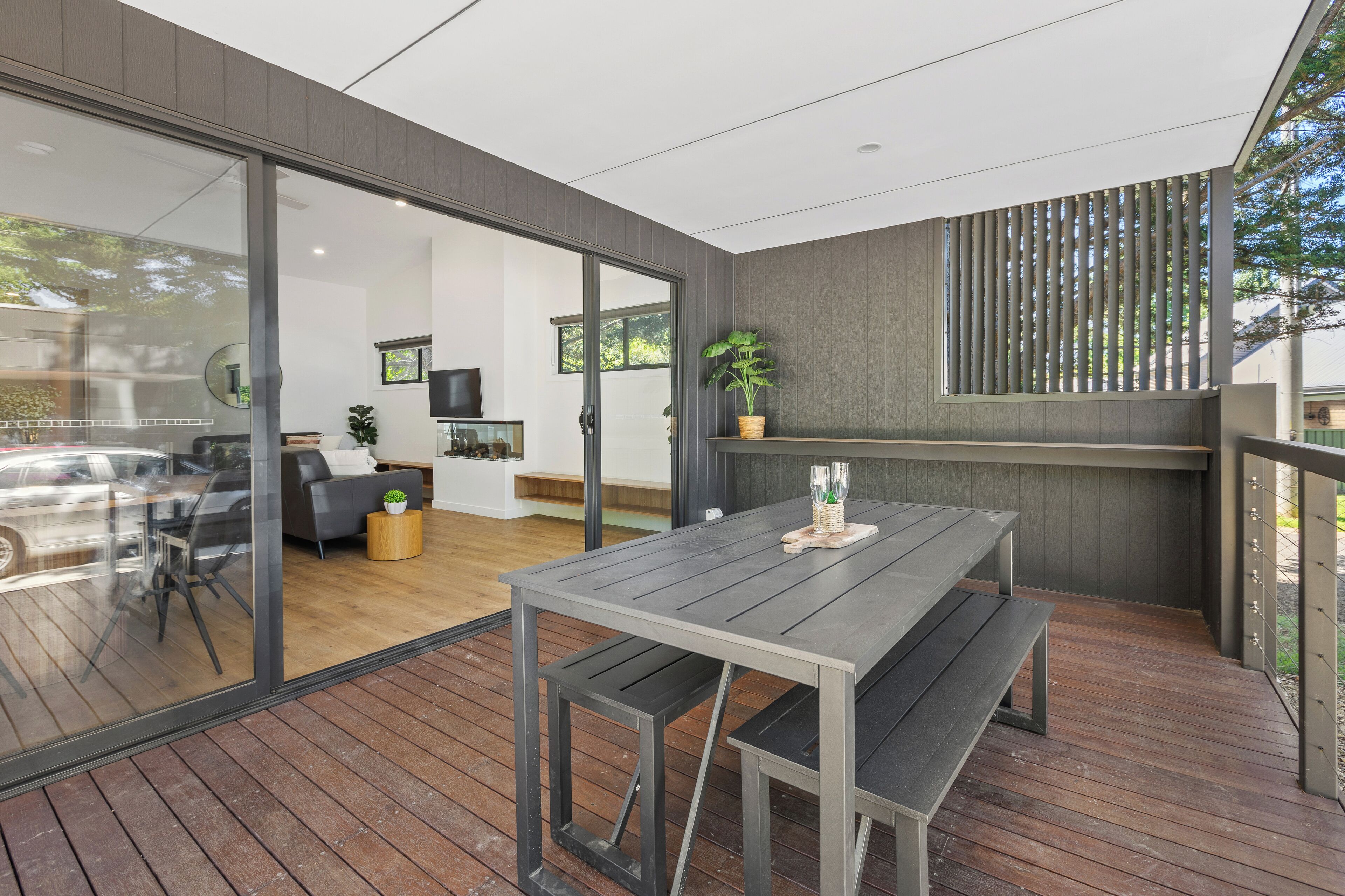 A modern outdoor dining area with a wooden table and benches, a glass door leading to a living room, and a vertical wooden slat design on the wall.
