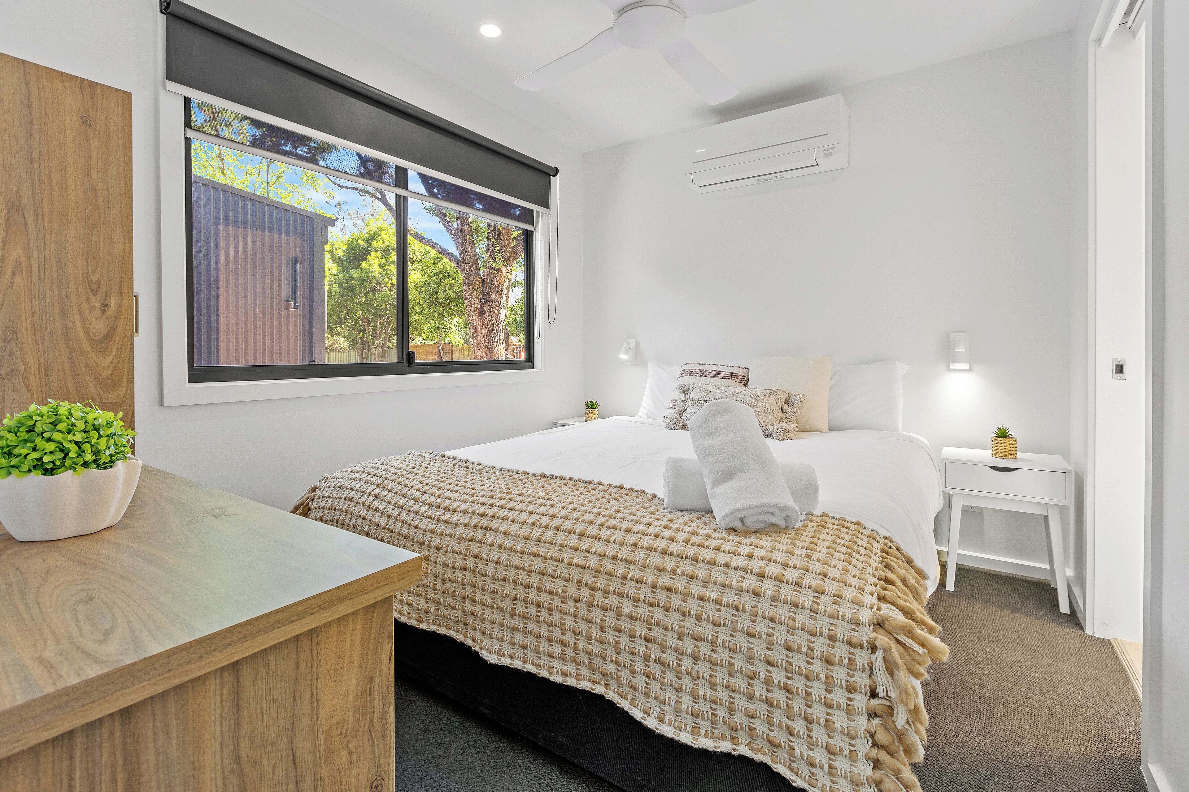 A modern bedroom with a large bed, a wooden desk, a small plant, and a window with a view of trees and a building.