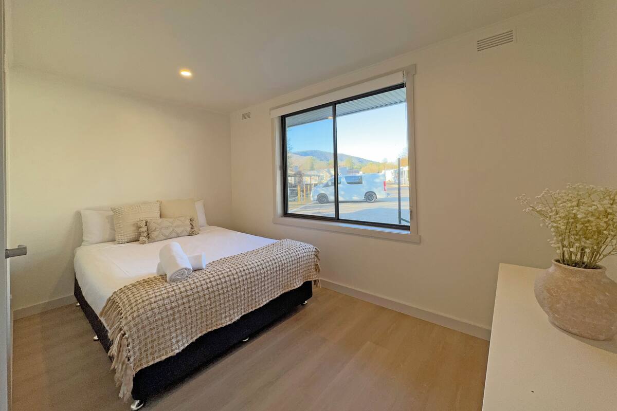 Quarto com cama, janela, mesa de cabeceira e um vaso com flores.