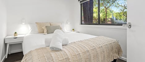 Et pænt opredt soveværelse med en stor seng, et sengebord med en lampe, og et vindue med udsigt til træer.