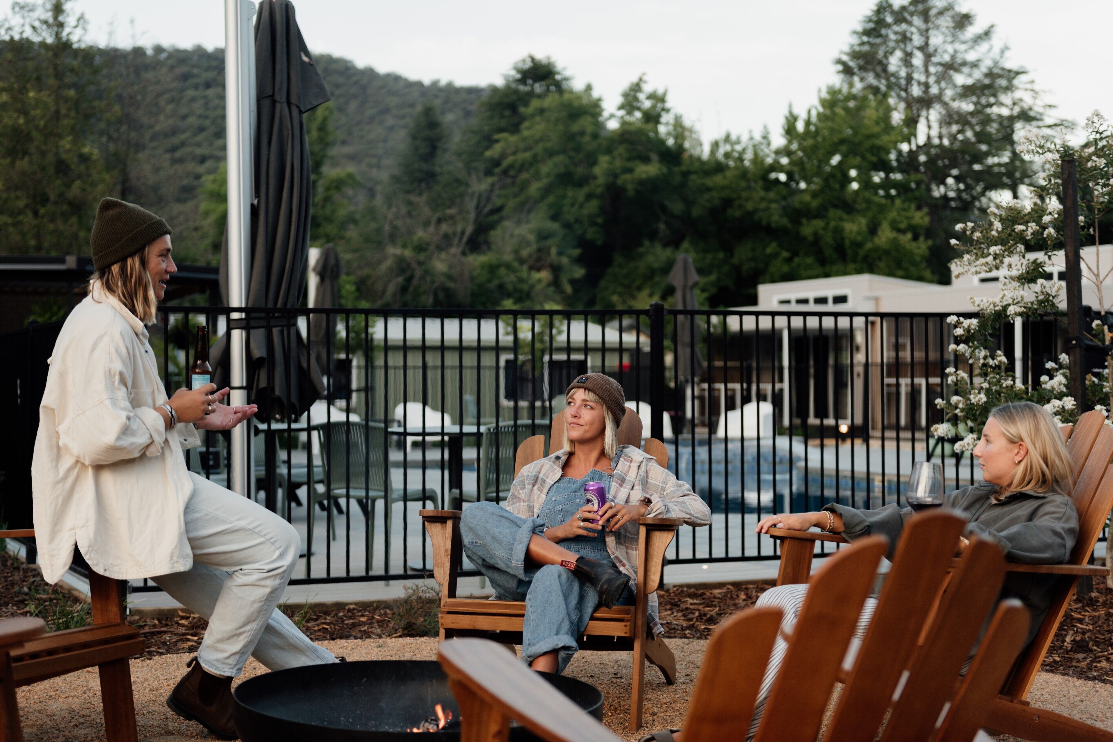 A group of people sitting on outdoor patio chairs near a pool, with a metal fence and greenery in the background.