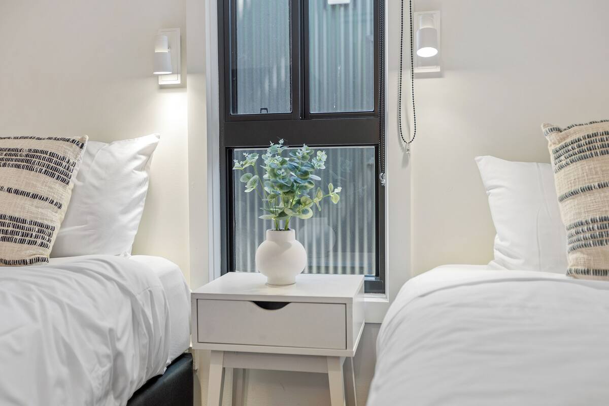 Quarto de hotel com duas camas, uma mesa de cabeceira branca com um vaso de flores e uma janela com persianas.