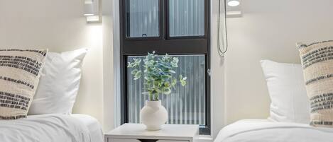 A hotel room with two beds, a white bedside table with a vase of flowers, and a window with blinds.