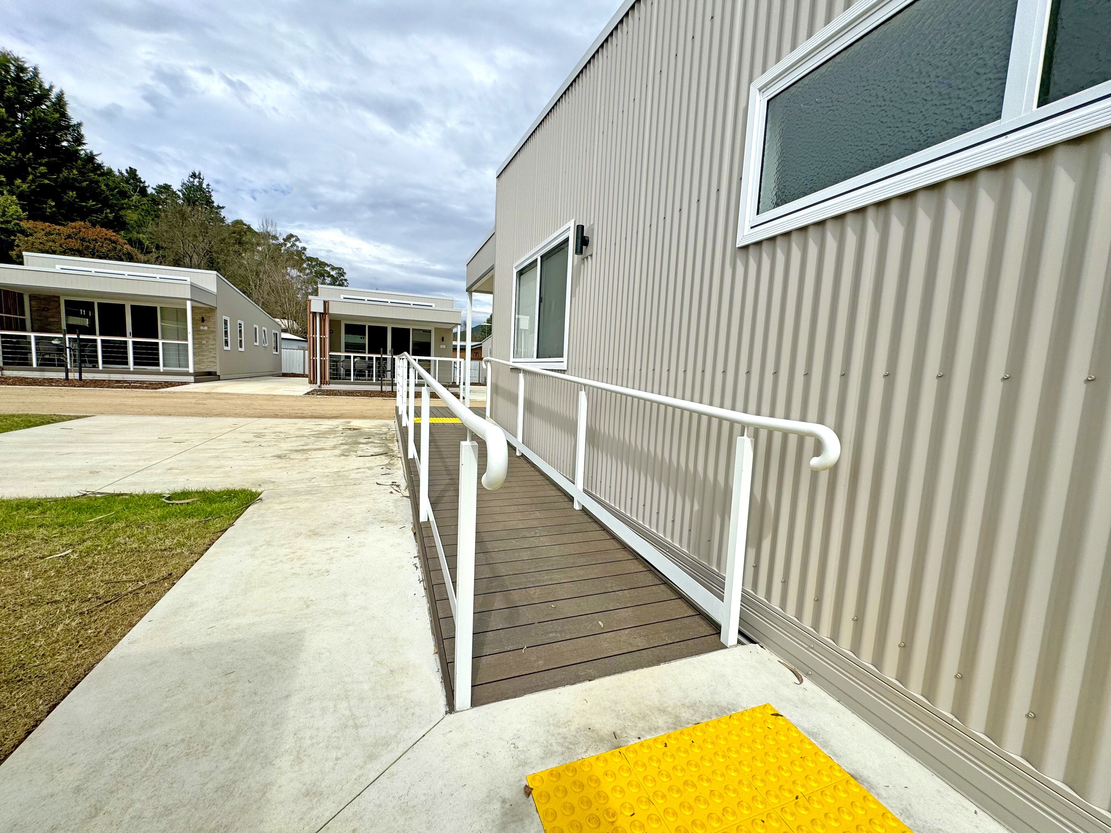A building with a ramp leading to a door, a yellow tactile paving at the bottom, and a white railing along the ramp.