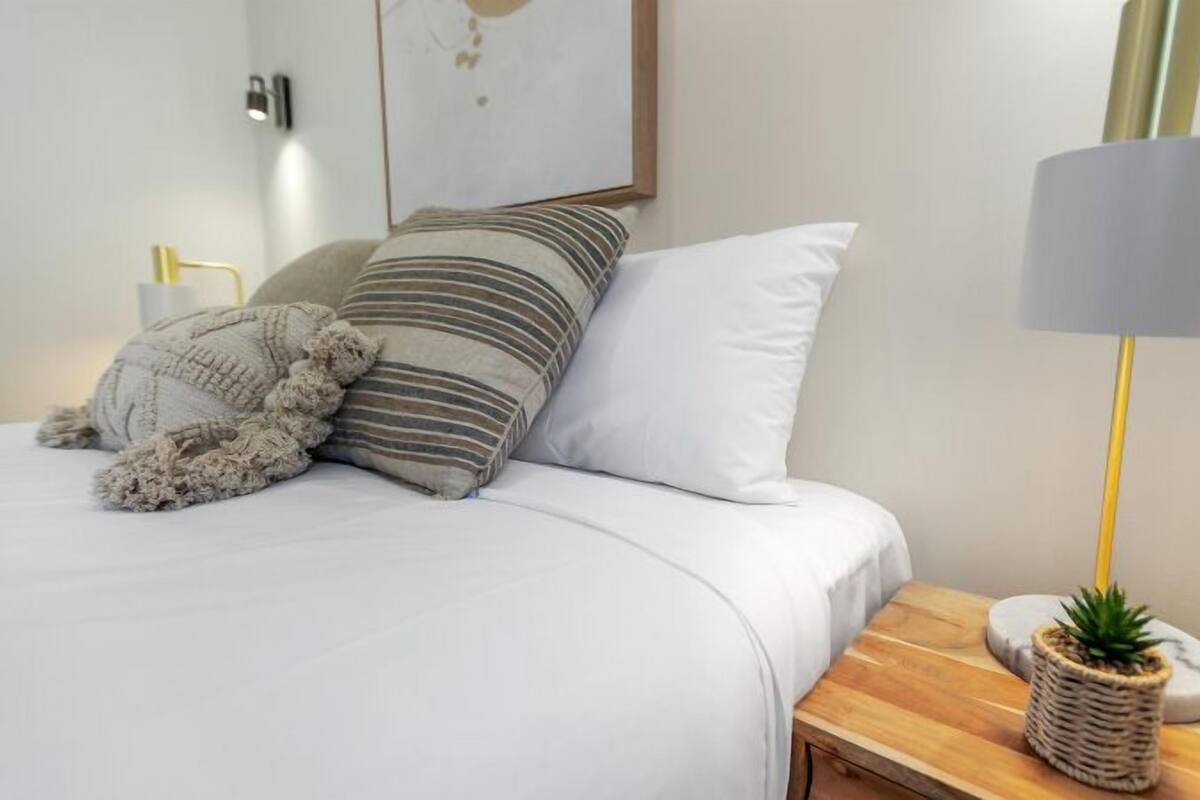 Uma cama bem arrumada com roupa de cama branca, almofadas listradas e lisas, um cobertor macio e uma mesa de cabeceira com um abajur e uma pequena planta.