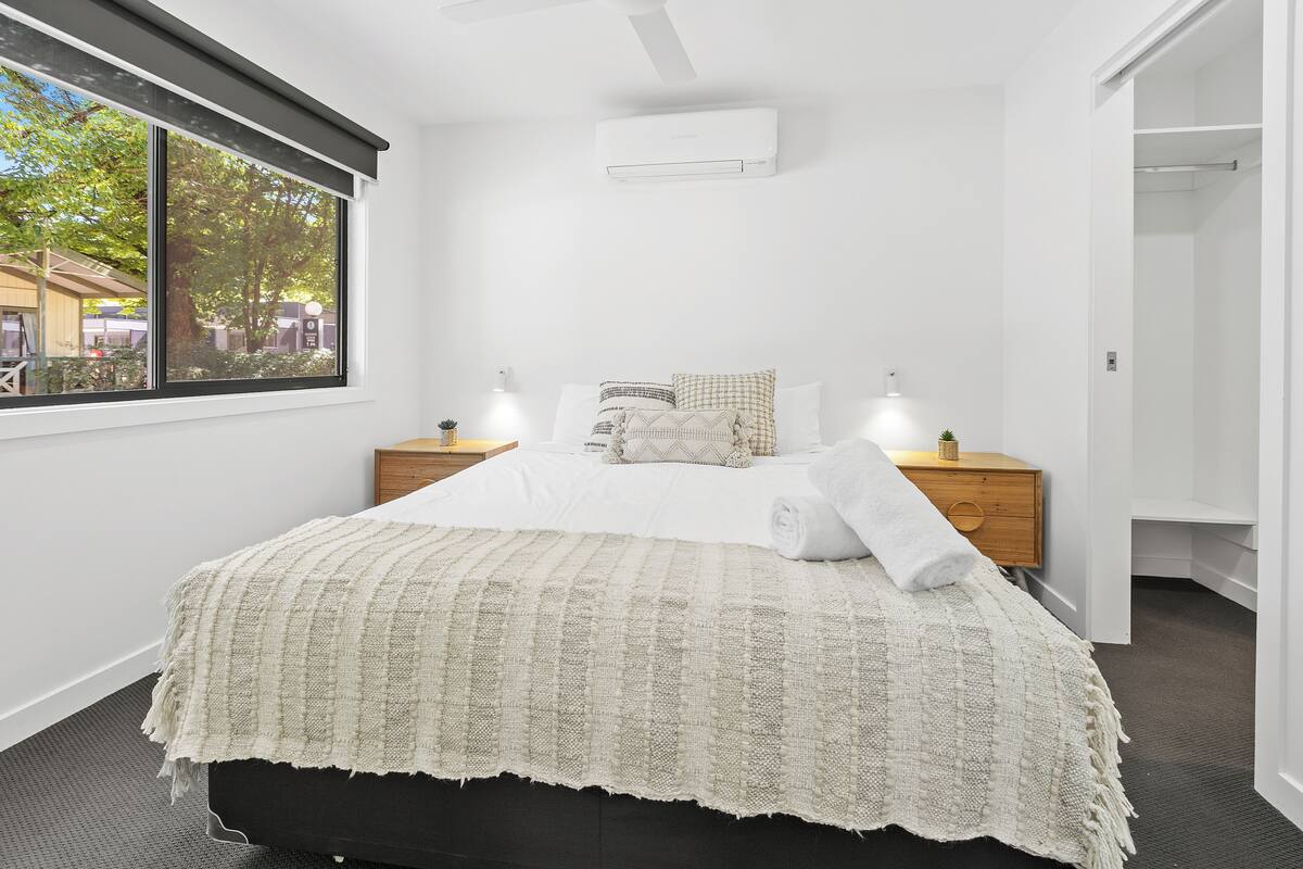 Quarto com uma janela grande, uma cama com cobertor branco, duas mesas de cabeceira com abajures e um criado-mudo de madeira.