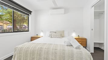 Quarto com uma janela grande, uma cama com cobertor branco, duas mesas de cabeceira com abajures e um criado-mudo de madeira.