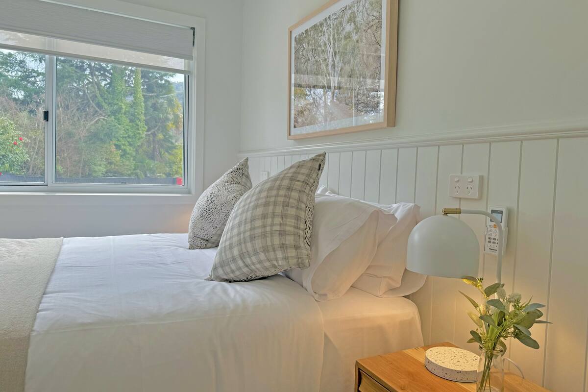 Cama bem arrumada com lençóis brancos e duas almofadas, mesa de cabeceira com abajur e um vaso de flores, e janela com vista para árvores.