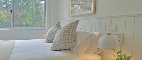 A neatly made bed with white linens and two pillows, a bedside table with a lamp and a vase of flowers, and a window with a view of trees.