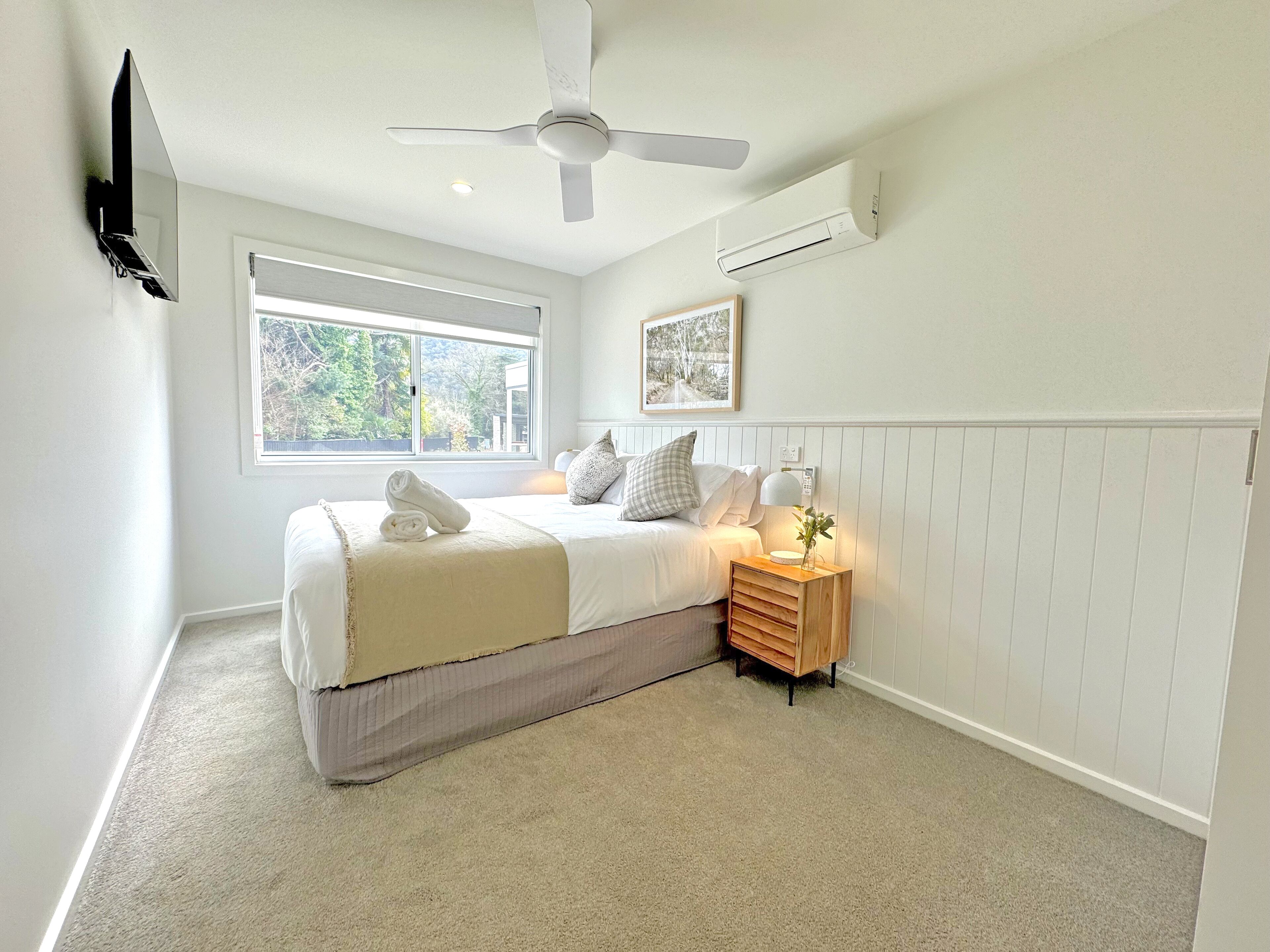 Un dormitorio con cama, mesita de noche con lámpara, ventilador de techo y ventana con vista a árboles.