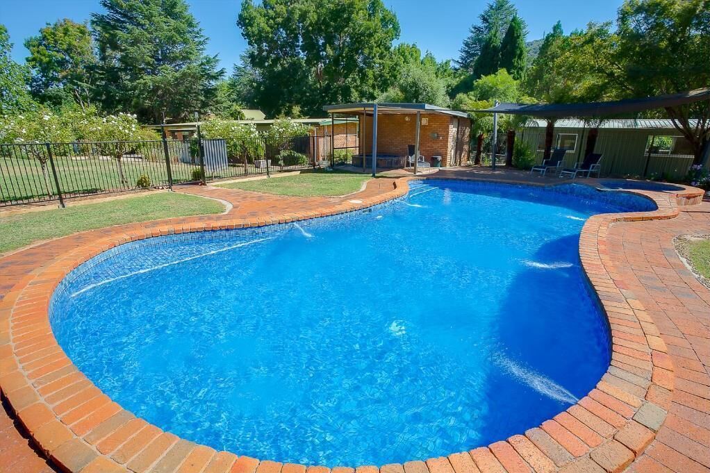 A swimming pool with a brick border, surrounded by a grassy area and trees.