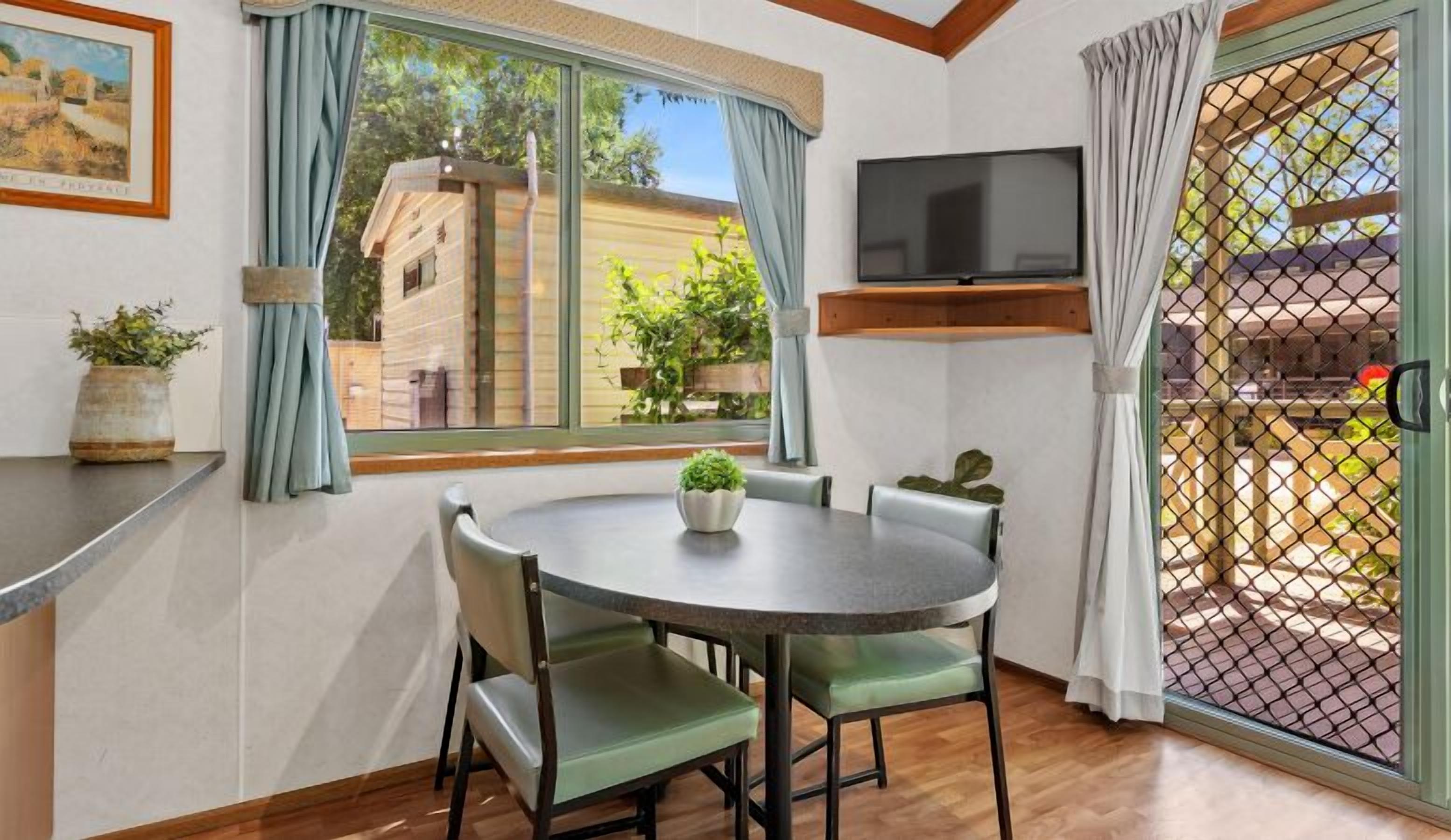 A dining area with a round table and chairs, a TV mounted on the wall, and a view of the outside through a window.