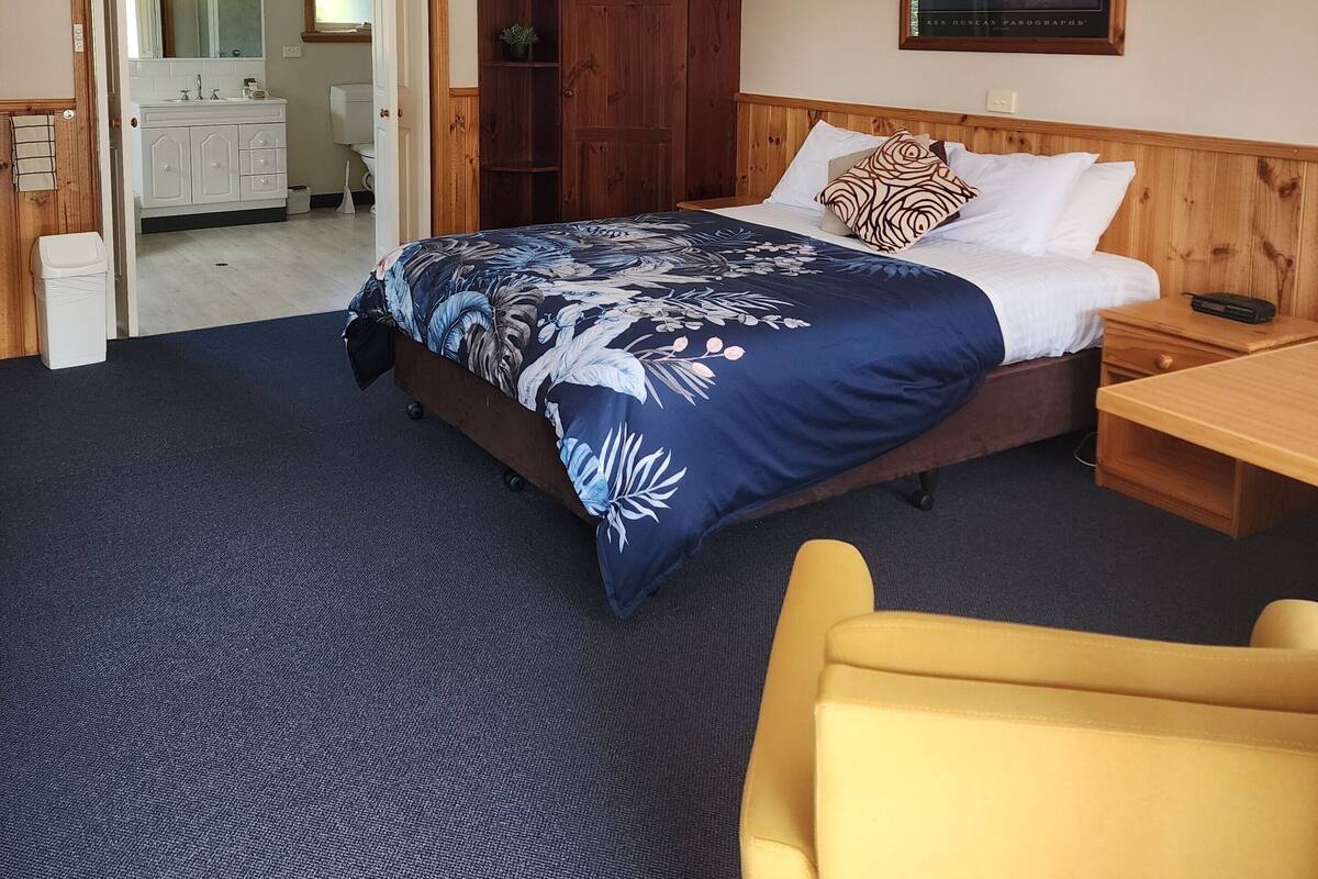 A hotel room with a bed, a wooden wardrobe, a nightstand, a yellow chair, and a framed picture on the wall.