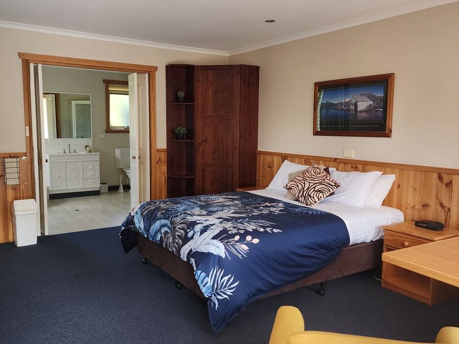A hotel room with a bed, a wooden wardrobe, a nightstand, a yellow chair, and a framed picture on the wall.