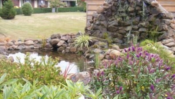 Una cabaña de madera rústica con un muro de piedra, un pequeño estanque y diversas plantas y flores en primer plano.