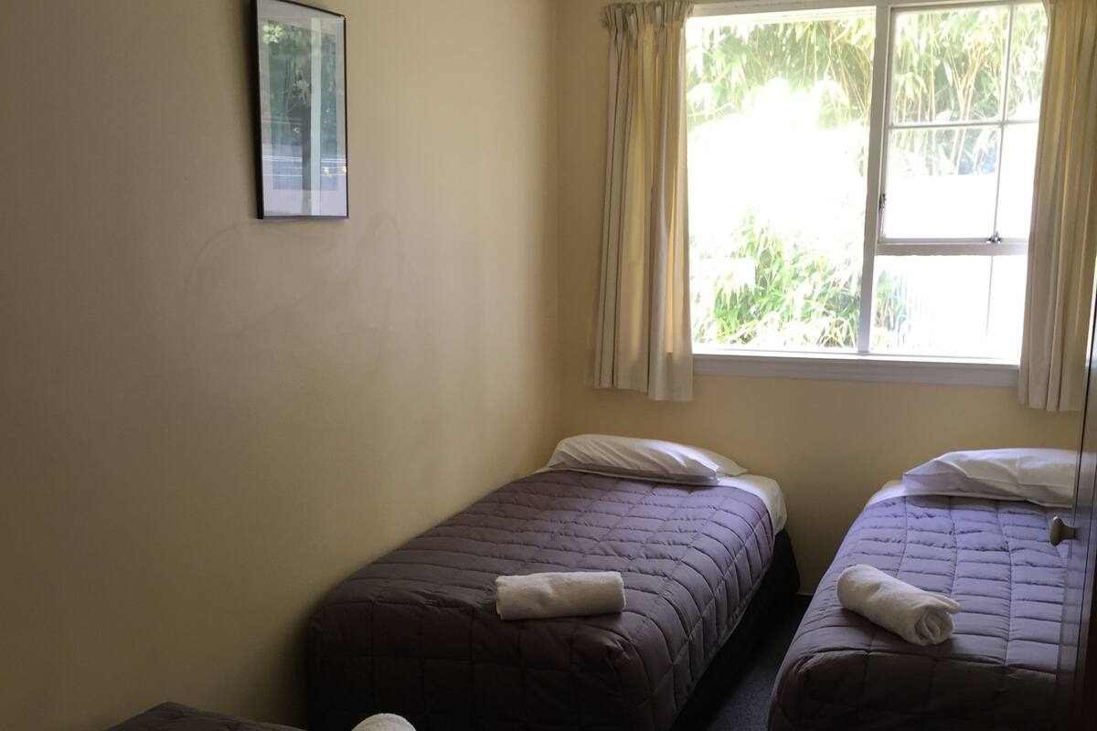 A room with three single beds, each with a rolled towel. There is a window with curtains, and a framed picture on the wall.