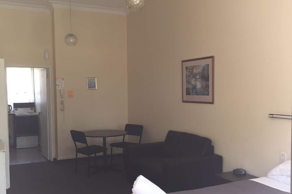 A hotel room with a bed, a small table with chairs, a painting on the wall, and a kitchenette in the background.