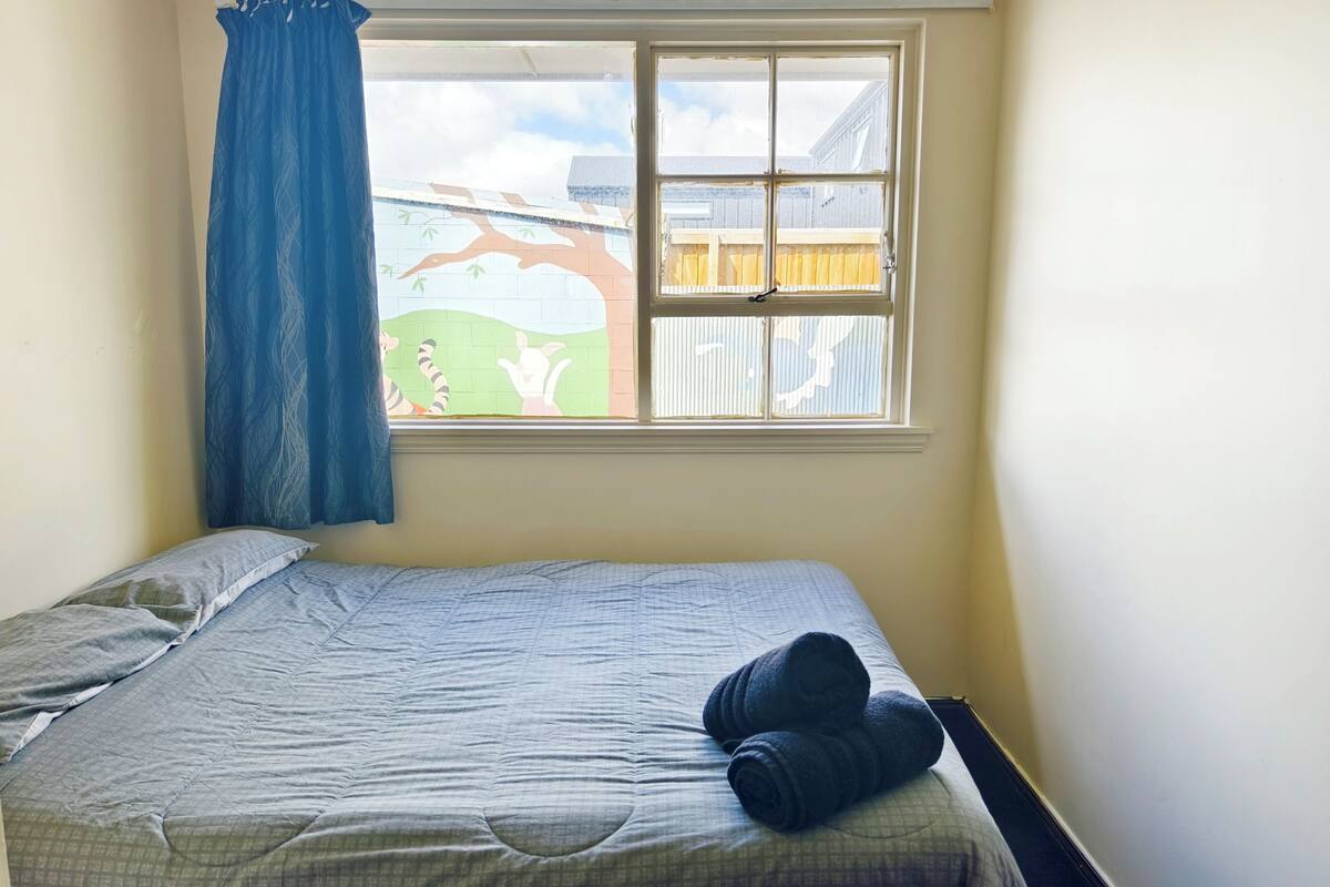 A single bed with a blue blanket and a pair of rolled towels, a window with a view of a fence and trees, and a blue curtain partially drawn.