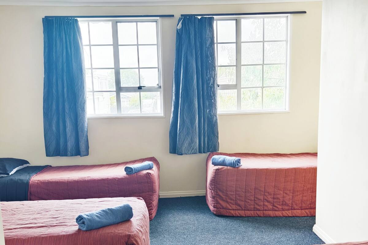 A room with three beds, each with a rolled towel, and a window with blue curtains.