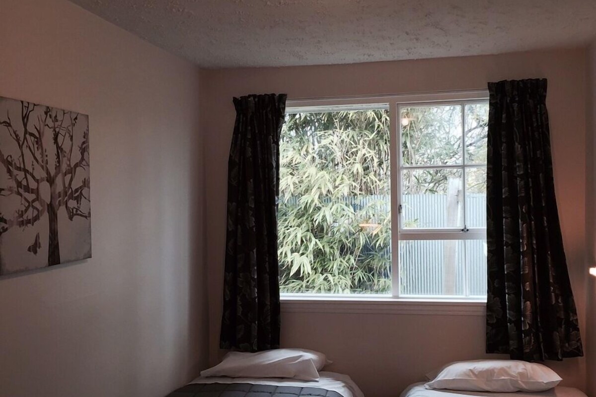 A room with two beds, a ceiling light, a window with curtains, and a tree painting on the wall.