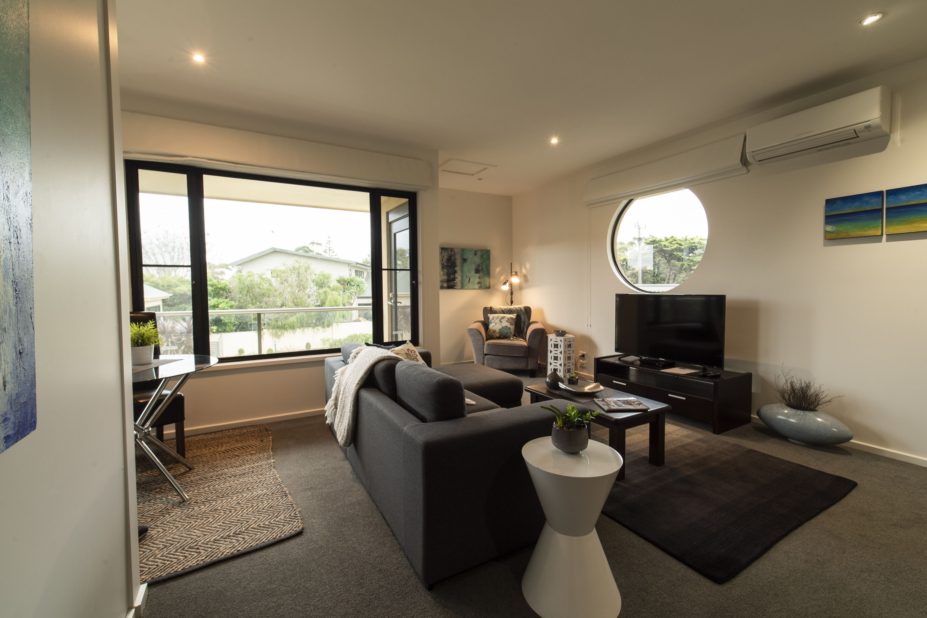 Ein modernes Wohnzimmer mit Sofa, Couchtisch und Fernseher. Ein großes Fenster bietet Blick ins Freie.