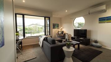 Ein modernes Wohnzimmer mit Sofa, Couchtisch und Fernseher. Ein großes Fenster bietet Blick ins Freie.
