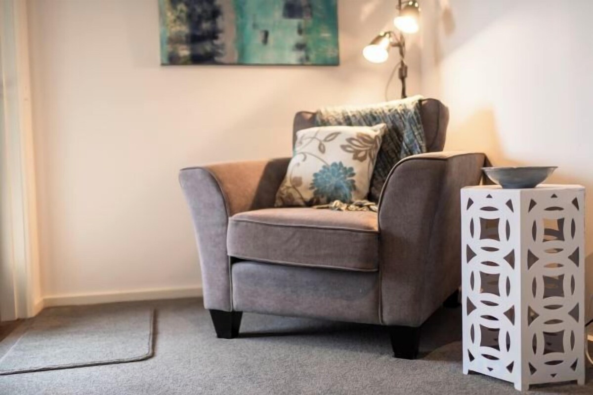 A grey sofa with floral cushions, a white side table with a bowl, and a painting on the wall.