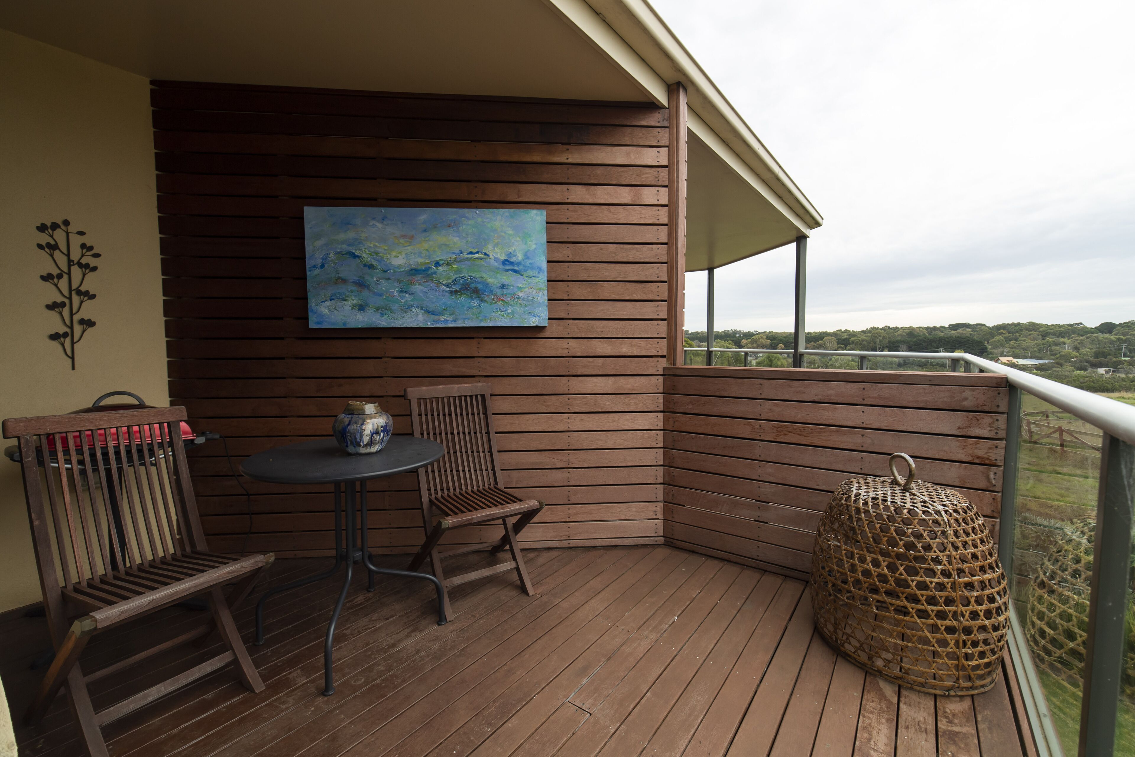 A wooden deck with a table and chairs, a painting on the wall, and a view of the landscape.