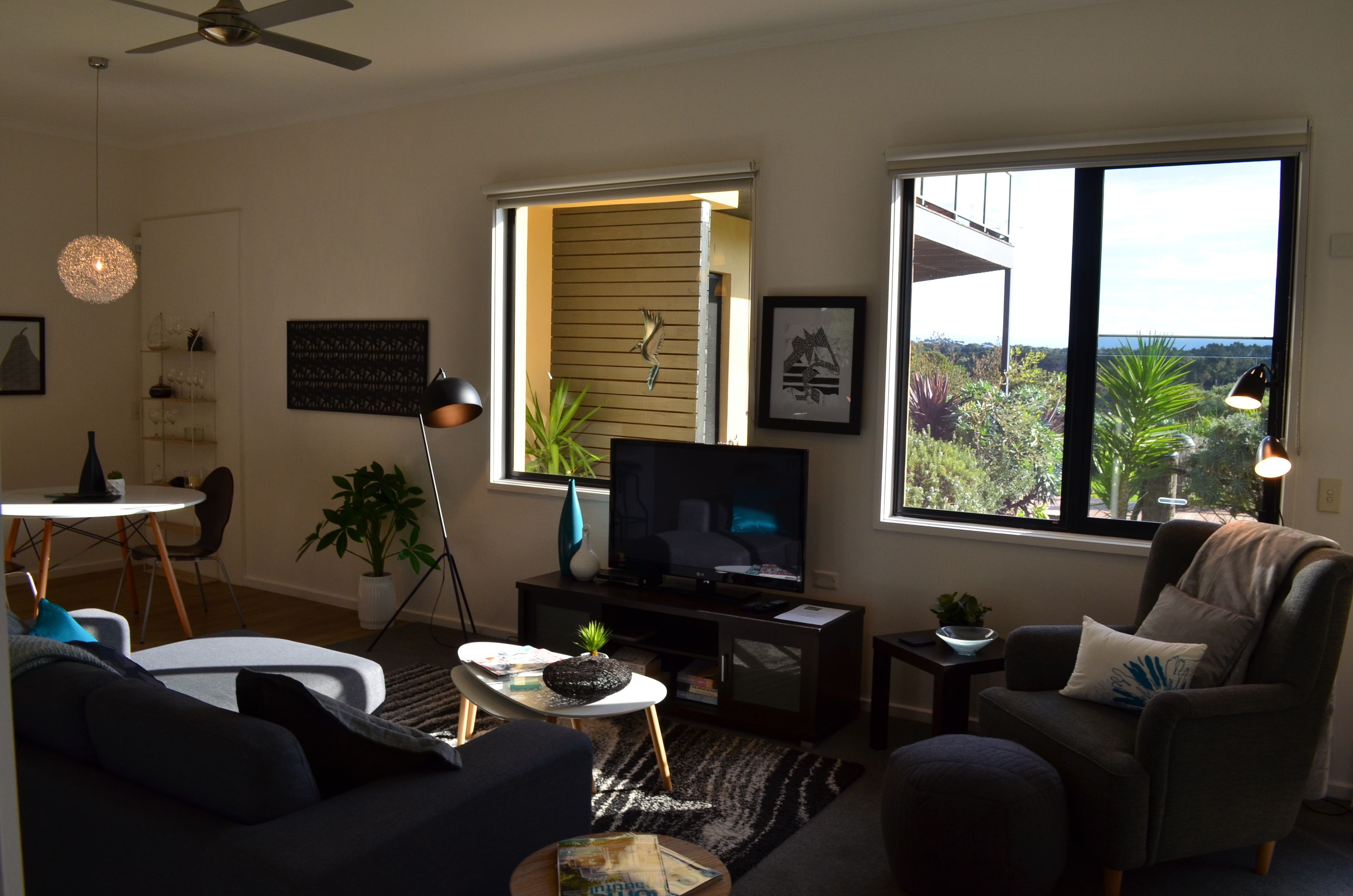 A living room with a television, a sofa, a chair, a small table, and a window with a view of greenery.
