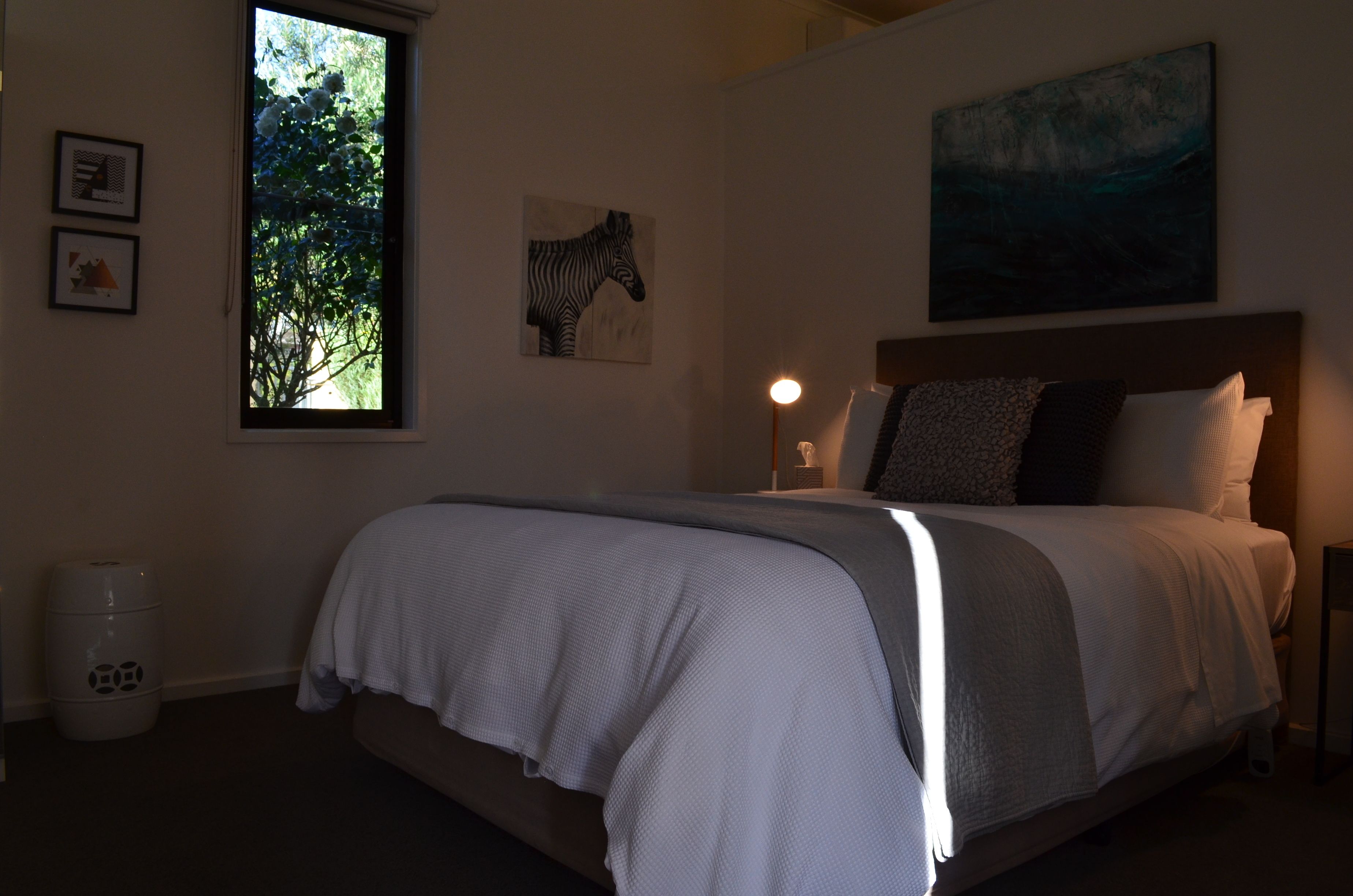 A bedroom with a large bed, a window with a view of trees, a zebra painting, and a landscape painting on the wall.
